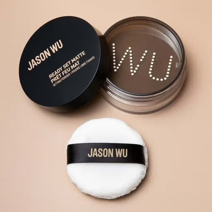 A container of JASON WU BEAUTY - READY SET MATTE 1 - TRANSLUCENT BANANA setting powder sits on a light brown textured surface. The cylindrical container has a black lid with the brand name in white capital letters and product details in both English and French, ensuring a flawless matte finish for any makeup look.