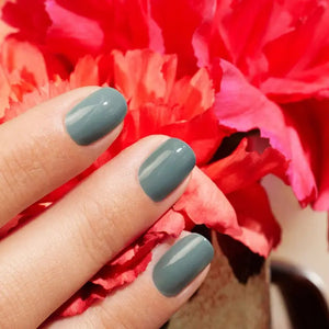 A hand with short, oval-shaped nails painted in MANUCURIST INC’s sophisticated GREEN™ Nail Polish in Sage—a chic gray vegan shade—is held in front of bright pink-red flower petals.