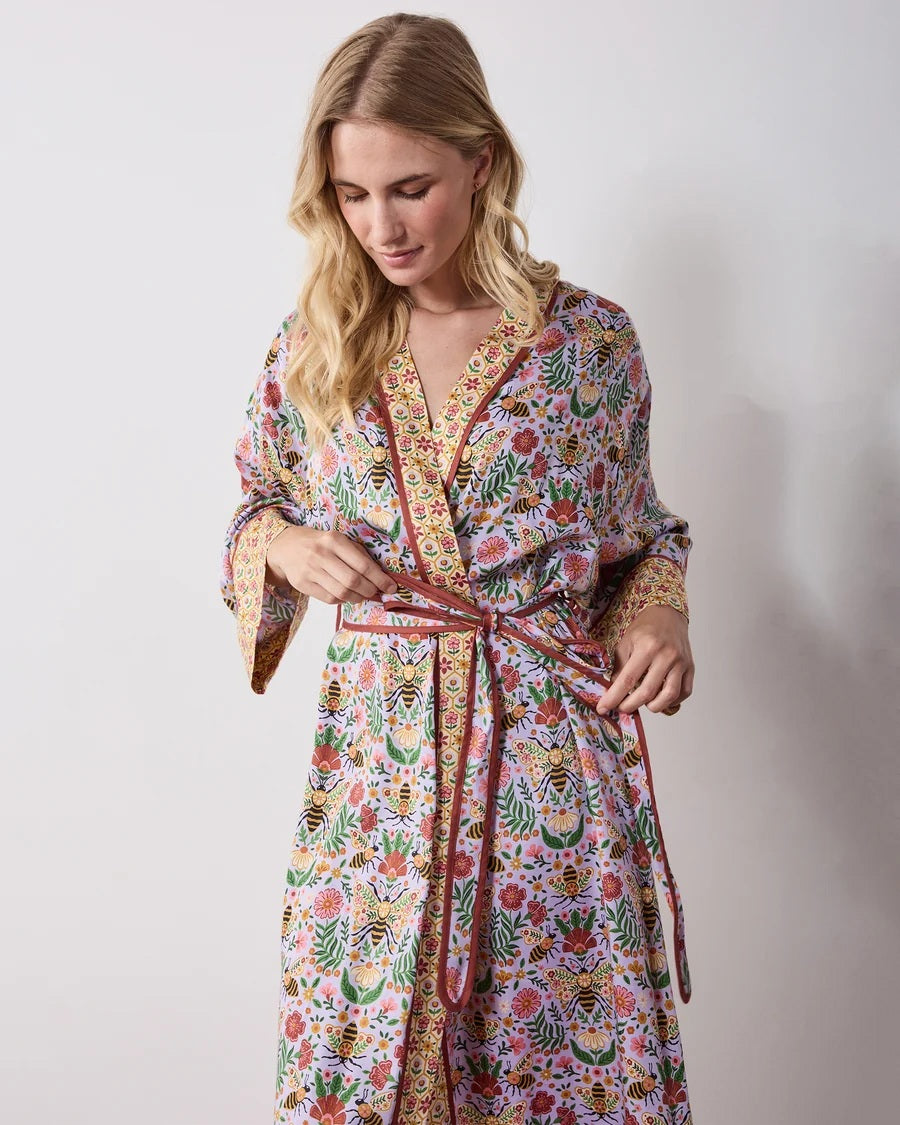 A woman wears a PRINTFRESH - BEE TAPESTRY ECO SATIN ROBE IN LAVENDER, featuring a lavender floral-print with a repeating pattern of bees, flowers, and vines with a contrasting yellow and red patterned trim.
