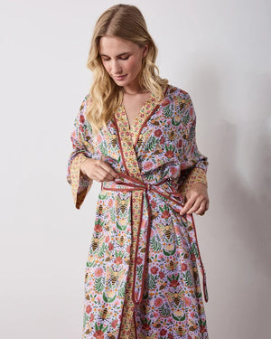 A woman wears a PRINTFRESH - BEE TAPESTRY ECO SATIN ROBE IN LAVENDER, featuring a lavender floral-print with a repeating pattern of bees, flowers, and vines with a contrasting yellow and red patterned trim.