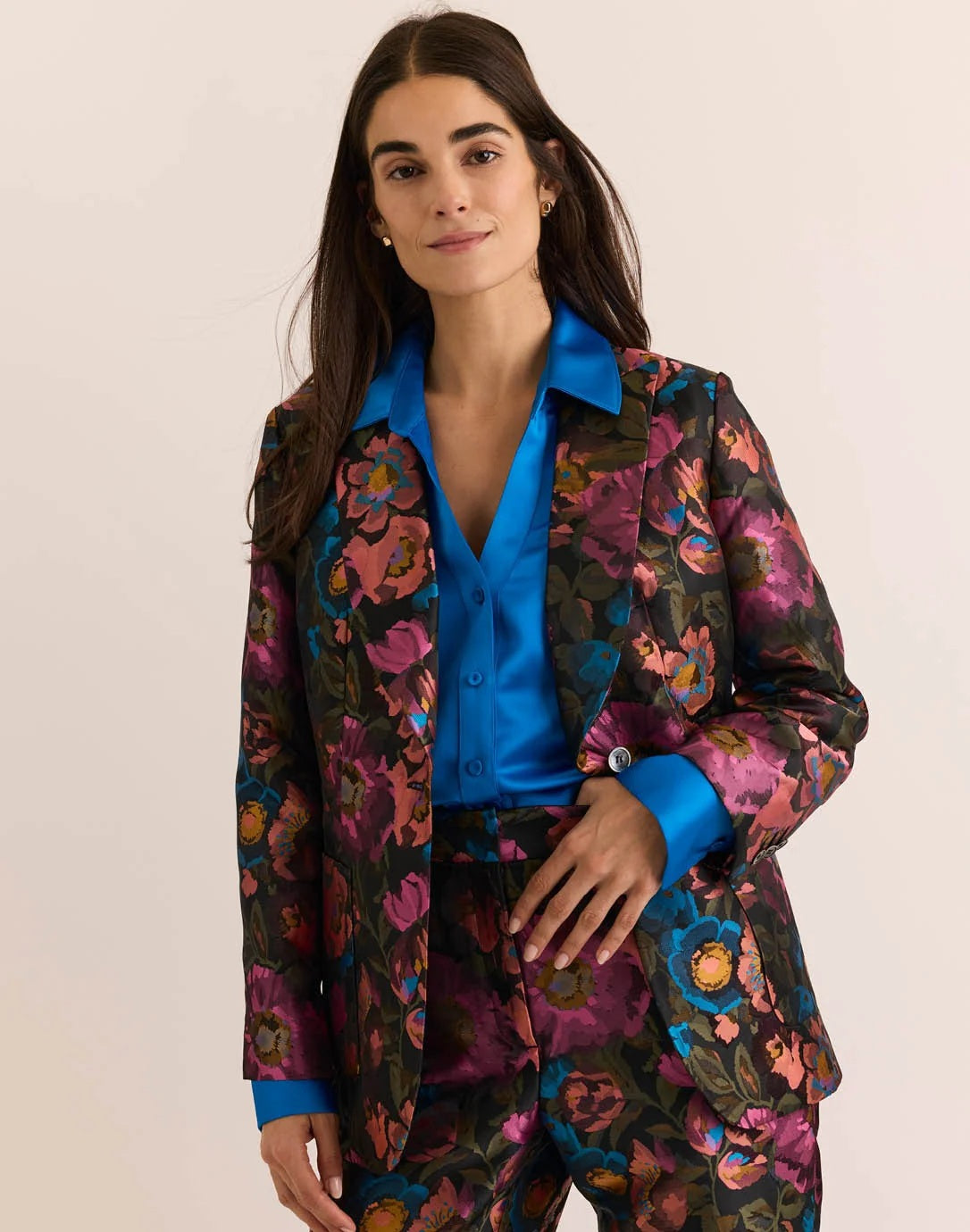 A woman with long brown hair is wearing the HINSON WU - CORINNE JACQUARD JACKET IN FLORAL BOUQUET, paired with a bright blue satin shirt underneath, standing against a plain light background.
