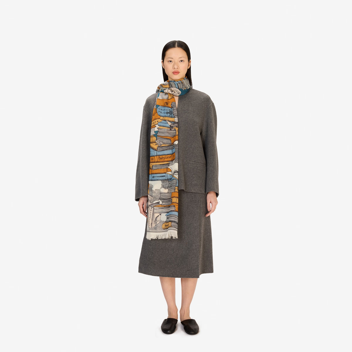 A woman stands facing forward, wearing a grey long-sleeve coat and skirt, black shoes, and the INOUI EDITIONS Stack of Books Wool Scarf in navy, a colorful, hand-printed accessory from INOUI, made in India, on a white background.