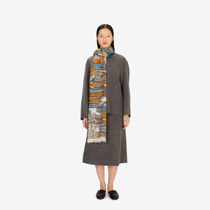 A woman stands facing forward, wearing a grey long-sleeve coat and skirt, black shoes, and the INOUI EDITIONS Stack of Books Wool Scarf in navy, a colorful, hand-printed accessory from INOUI, made in India, on a white background.