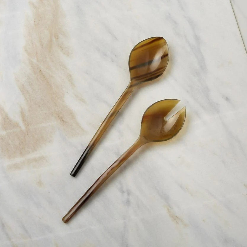 Two brownish-colored utensils from the BE HOME, INC HORN THIN SERVING SET lay on a light marble surface. One is a regular spoon, and the other is an artisan-crafted spork with a single notch. Both have a glossy finish and appear to be made of ethically salvaged material resembling horn.