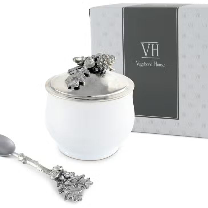 The ACORN OAK LEAF SUGAR BOWL by VAGABOND HOUSE is a small fine porcelain bowl with a silver lid adorned with intricate decorative elements, including acorns and oak leaves. The lid&#39;s handle is an ornate oak leaf design, enhancing the bowl&#39;s elegant and detailed appearance, reminiscent of a luxury grade pewter Antler Creamer Set.