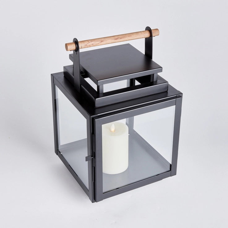 The NAPA HOME AND GARDEN - ADWIN OUTDOOR LANTERN is a black metal lantern with clear glass panels and a unique design, featuring a teak handle. It holds an unlit white candle inside, making it ideal for outdoor use with its contemporary and minimalist style.
