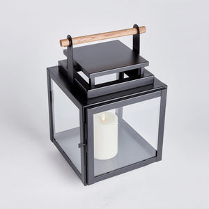 The NAPA HOME AND GARDEN - ADWIN OUTDOOR LANTERN is a black metal lantern with clear glass panels and a unique design, featuring a teak handle. It holds an unlit white candle inside, making it ideal for outdoor use with its contemporary and minimalist style.