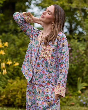 A person wearing the PRINTFRESH - BEE TAPESTRY ECO SATIN SOHO SET IN LAVENDER, featuring vintage-inspired bee and floral vine prints, holding a flower behind their back in a garden.