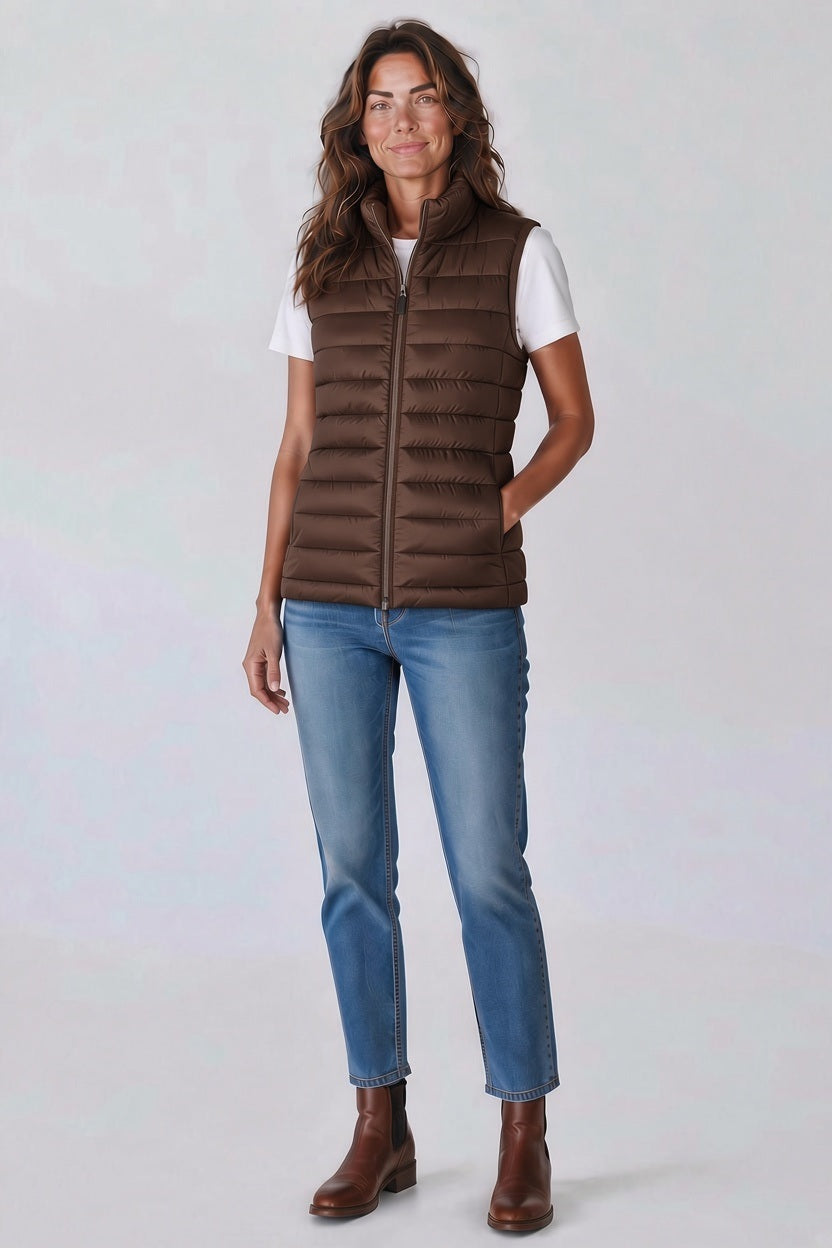 A woman wears a brown SAVE THE DUCK - LYNN PUFFER VEST IN NOISETTE BROWN over a white t-shirt, blue jeans, and brown ankle boots.