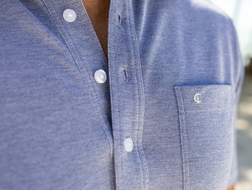 A close-up of a person wearing the CRIQUET - Oxford Pique Players Shirt in First Mate, featuring white buttons, a left chest pocket with a small embroidered logo. The person's face is not visible.