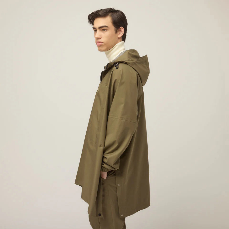A person models the TILLEY - PACKABLE HOODED PONCHO in olive green, layered over a white turtleneck, standing against a plain light background and gazing to the side with a neutral expression. Brand: TILLEY.