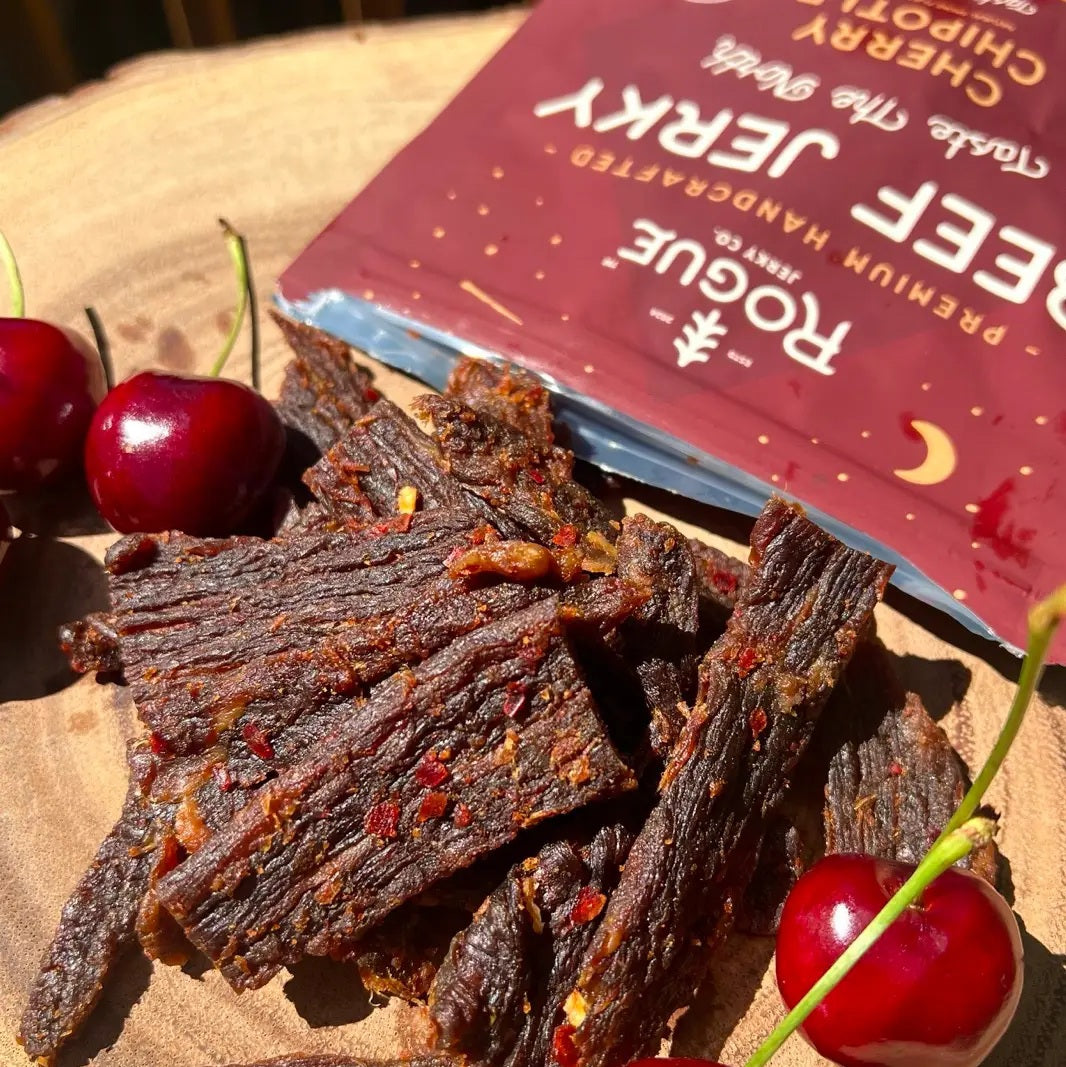 A maroon package of ROGUE JERKY CO. ROGUE JERKY - CHERRY CHIPOTLE BEEF JERKY, decorated with tree and mountain illustrations. The packaging highlights “Premium Handcrafted” and tasting notes of “Fruity, Warm, Vibrant.” This high-protein gourmet snack comes in a 2-ounce net weight.