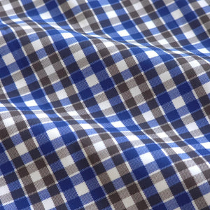 A man wearing sunglasses and the COLLARS AND CO - Semi Spread Collar Apex Performance Dress Shirt in Royal and Brown Tattersall stands confidently on a sunny, palm-lined street.