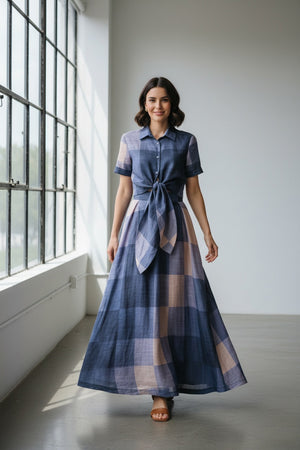 A woman wearing oversized orange sunglasses and the LAVI - MENTA Maxi Skirt in Quadri by LAVI, a long blue and beige plaid skirt with a tied waist and flowing maxi silhouette, stands against a plain white background.
