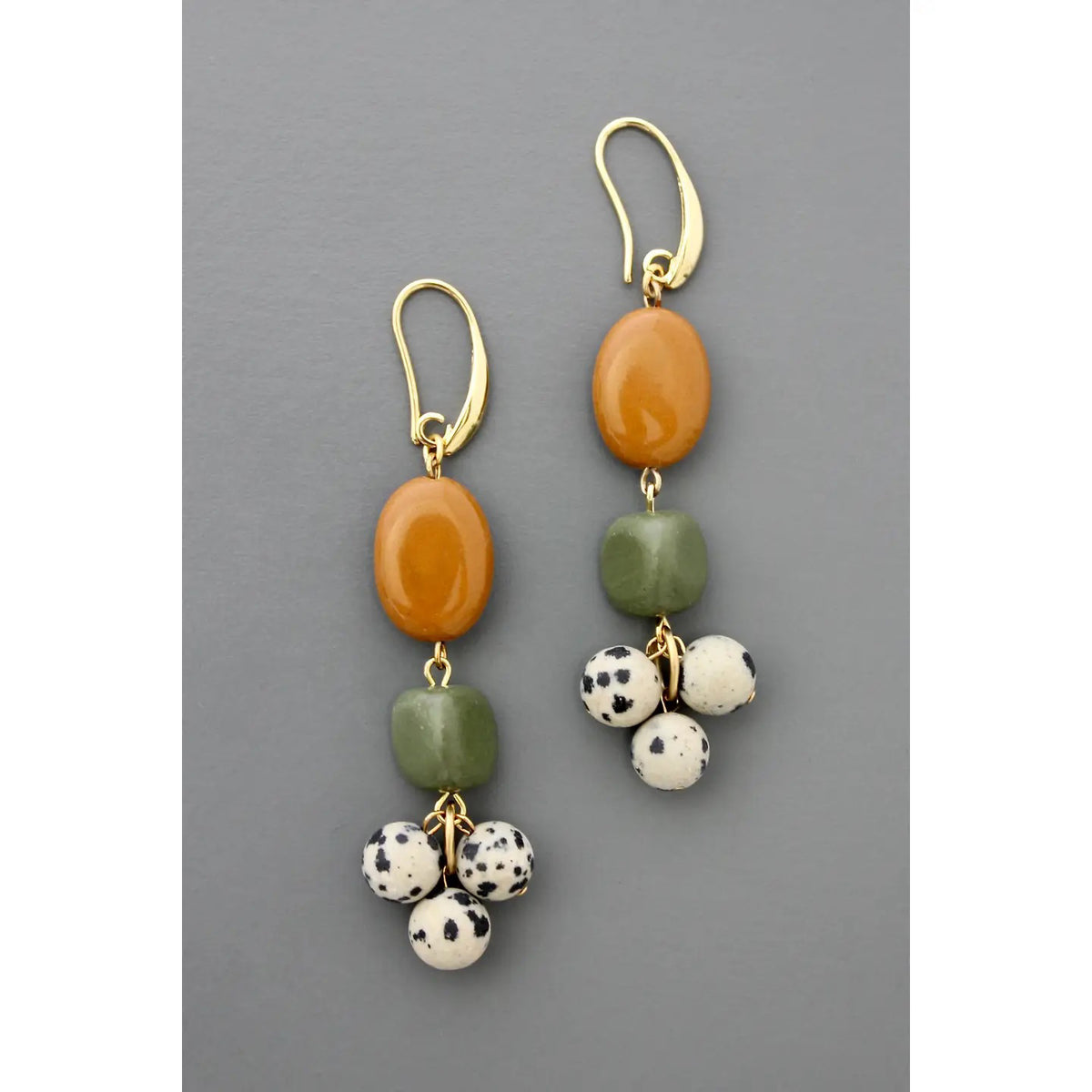 DAVID AUBREY JASPER SERPENTINE DALMATION CLUSTER EARRINGS by DAVID AUBREY feature gold-plated brass, an oval amber bead, a square green bead, and three small Dalmatian-spotted white beads against a gray background.
