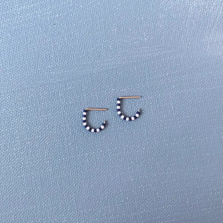 A close-up of a person’s ear wearing ALICE RISE - TINY HUGGIE EARRINGS IN NAVY, the handmade jewelry partly hidden by green leaves as natural light from a window highlights the unique ALICE RISE design.