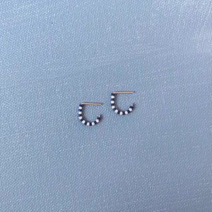 A close-up of a person’s ear wearing ALICE RISE - TINY HUGGIE EARRINGS IN NAVY, the handmade jewelry partly hidden by green leaves as natural light from a window highlights the unique ALICE RISE design.