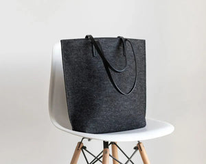 A person in a beige trench coat holds the WOOLBERRY WOOL FELT LONG TOTE BAG with black handles, standing against a plain, light-colored background.