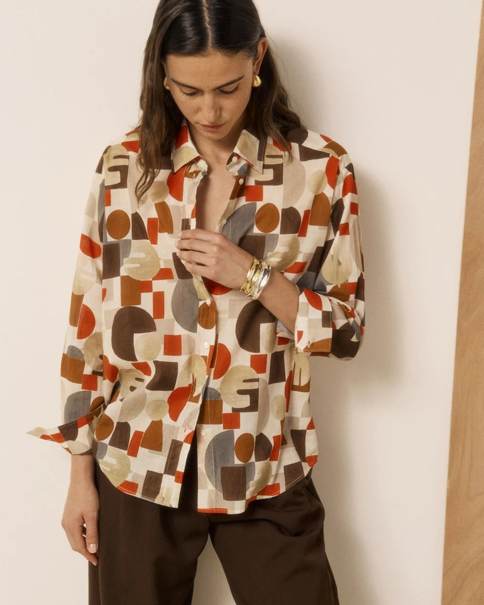 A woman with wavy brown hair stands barefoot indoors, wearing brown wide-leg pants and the HARTFORD HATFORD - CINEMA SHIRT GEOMETRIC PRINT—a loose-fit shirt featuring an abstract orange, beige, and gray pattern—as she gazes out a large window.