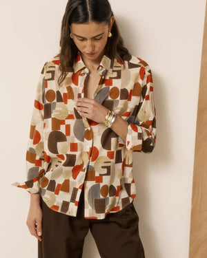 A woman with wavy brown hair stands barefoot indoors, wearing brown wide-leg pants and the HARTFORD HATFORD - CINEMA SHIRT GEOMETRIC PRINT—a loose-fit shirt featuring an abstract orange, beige, and gray pattern—as she gazes out a large window.
