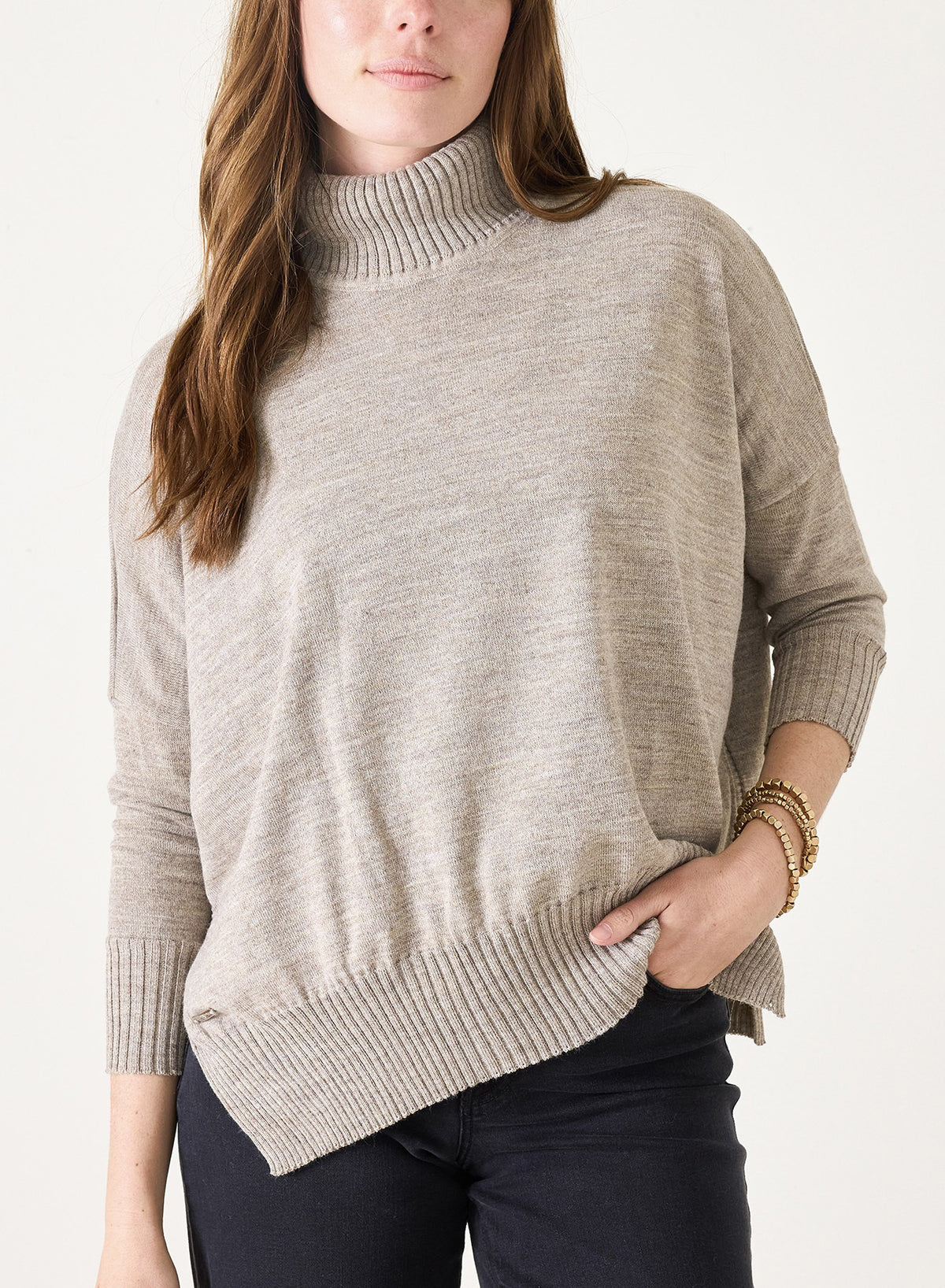 A woman wearing the MERSEA Catalina Turtleneck in Driftwood, paired with dark pants and gold bracelets, stands with one hand in her pocket against a plain background.