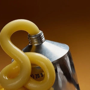 A metallic tube labeled “HONEY DEPARTMENT- THICK HONEY TUBE” stands upright against a brown gradient background. The design features yellow dots and bold black text, resembling a toothpaste or paint tube—making it perfect for dispensing thick honey with ease from HONEY DEPARTMENT.