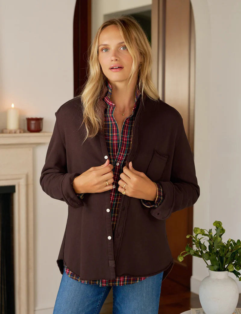 A woman with long blonde hair wears the FRANK AND EILEEN - EILEEN Relaxed Button Up Shirt in Triple Fleece Coco, by FRANK &amp; EILEEN. Indoors, she looks down and adjusts her sleeve in a cozy, warmly lit room.