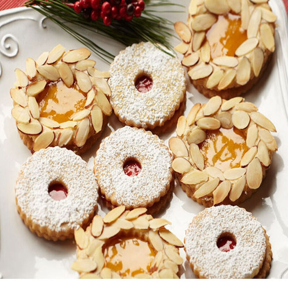 A plate of festive cookies, some topped with sliced almonds and filled with STONEWALL KITCHEN - APRICOT JAM for a juicy sweetness, others dusted with powdered sugar and filled with red jam. Pine and berries decorate the edge of the plate.