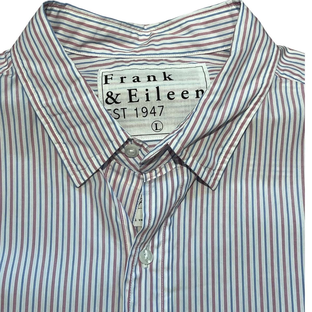 A man with short gray hair and a beard sits on a park bench wearing the FRANK & EILEEN Finbar men's button-up shirt in pink and blue multi stripe, blue jeans, and white sneakers. Behind him, people walk along a flower-lined path among trees.