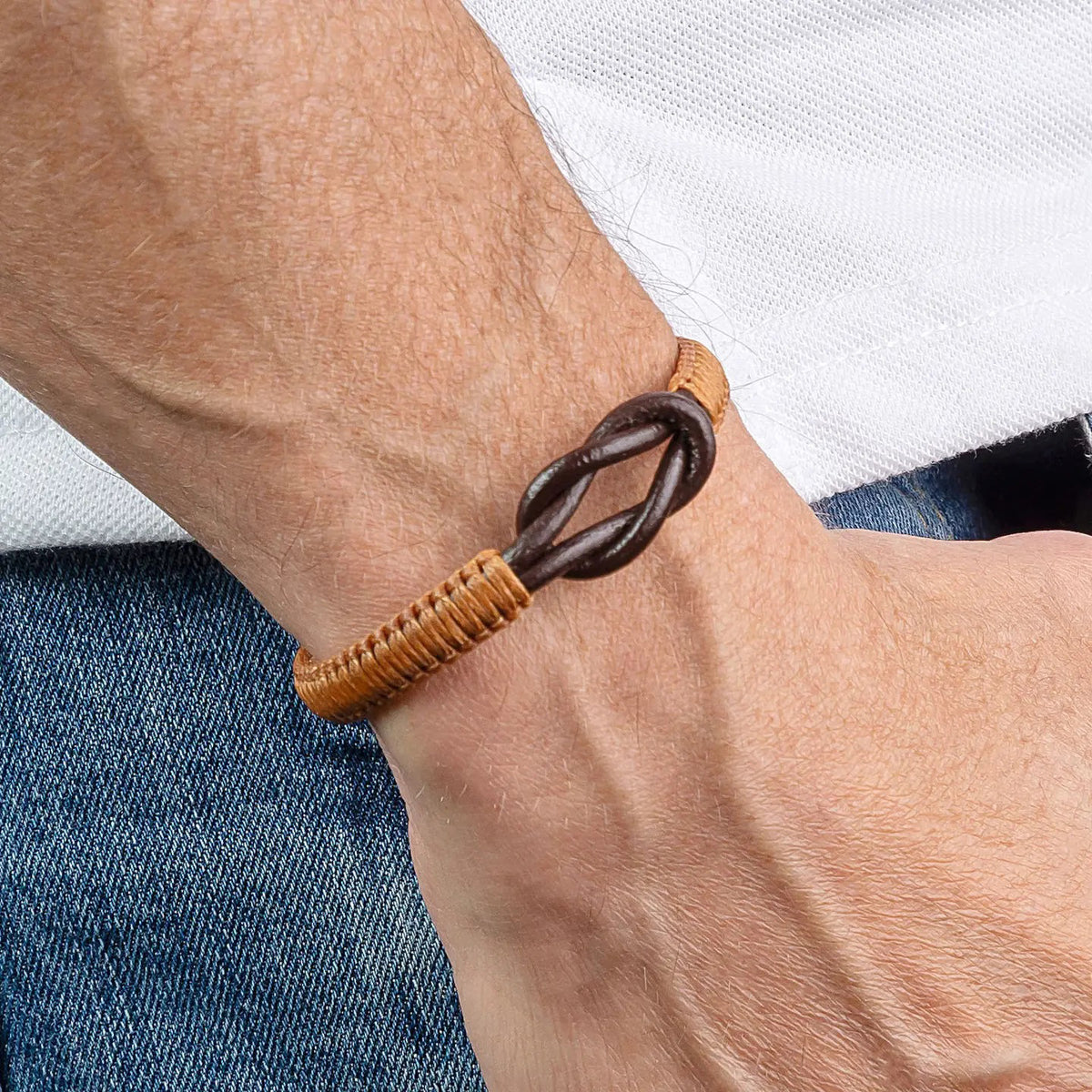 The MAKARLA Men's Infinity Knot Leather Bracelet in chocolate features a handmade design with genuine leather and an adjustable sliding knot, displayed on a white cylindrical stand and labeled by color.