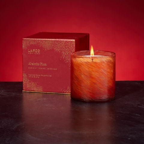 A glowing, orange-colored candle made from soy wax is placed next to a red box labeled &quot;LAFCO - ABSINTHE PLUM HOLIDAY CANDLE 15.5OZ.&quot; The scent of black plum and sandalwood permeates the air. The candle&#39;s warm, inviting glow is beautifully enhanced by the rich red background.