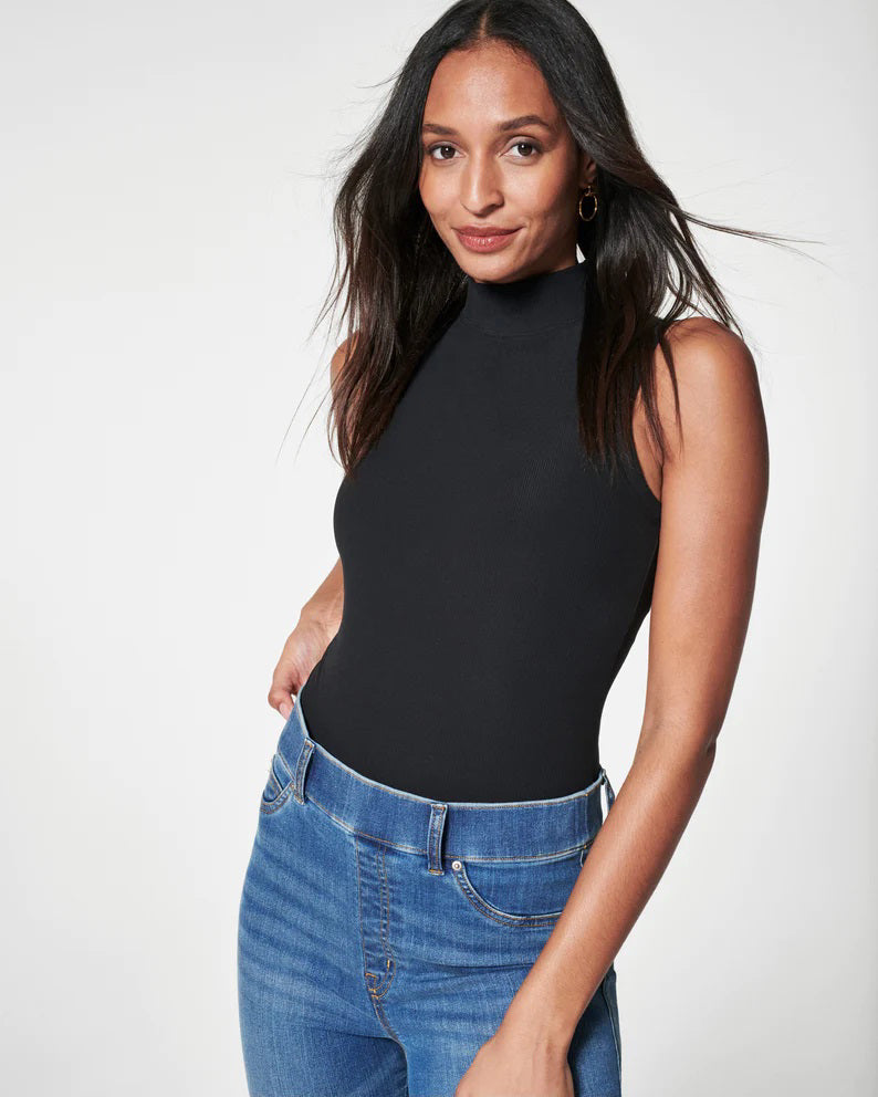 A woman poses against a white background in the SPANX jeannish high-waisted blue flare jeans made from premium stretch denim with hidden core shaping technology, styled with a black faux leather jacket, black top, and pointed-toe shoes.