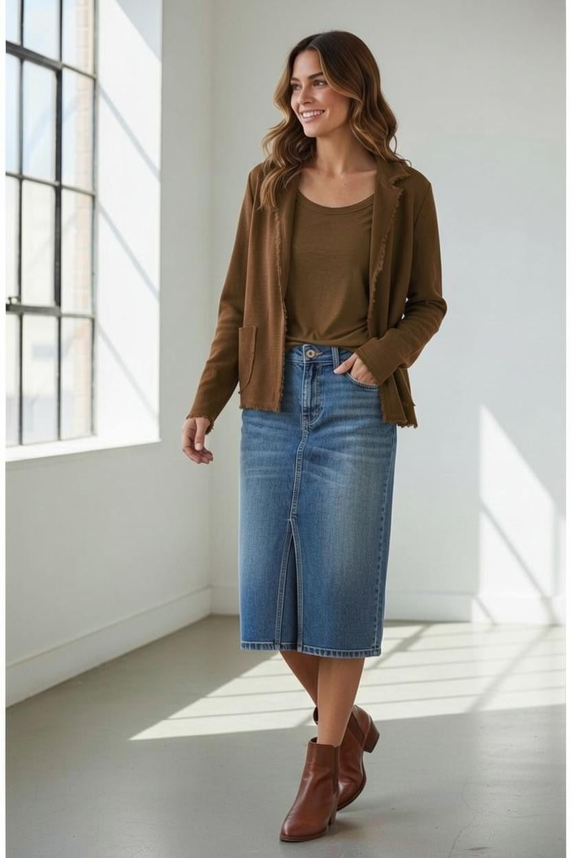 A woman stands indoors near large windows, smiling and looking to the side. She is wearing the BASE MILANO - COTTON KNIT CARDIGAN IN BROWN layered effortlessly over a brown top, paired with a knee-length denim skirt and brown ankle boots—an ideal look for transitional weather.