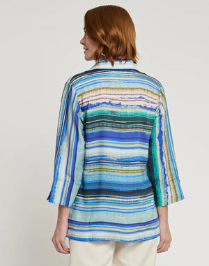 A person with shoulder-length auburn hair is wearing the HINSON WU - VICKY 3/4 Sleeve Tencel Textured Stripe Print Top, which is a loose-fitting shirt featuring vibrant horizontal stripes in blue, green, and white. They are facing slightly left, highlighting the side of the outfit against a plain light gray background.