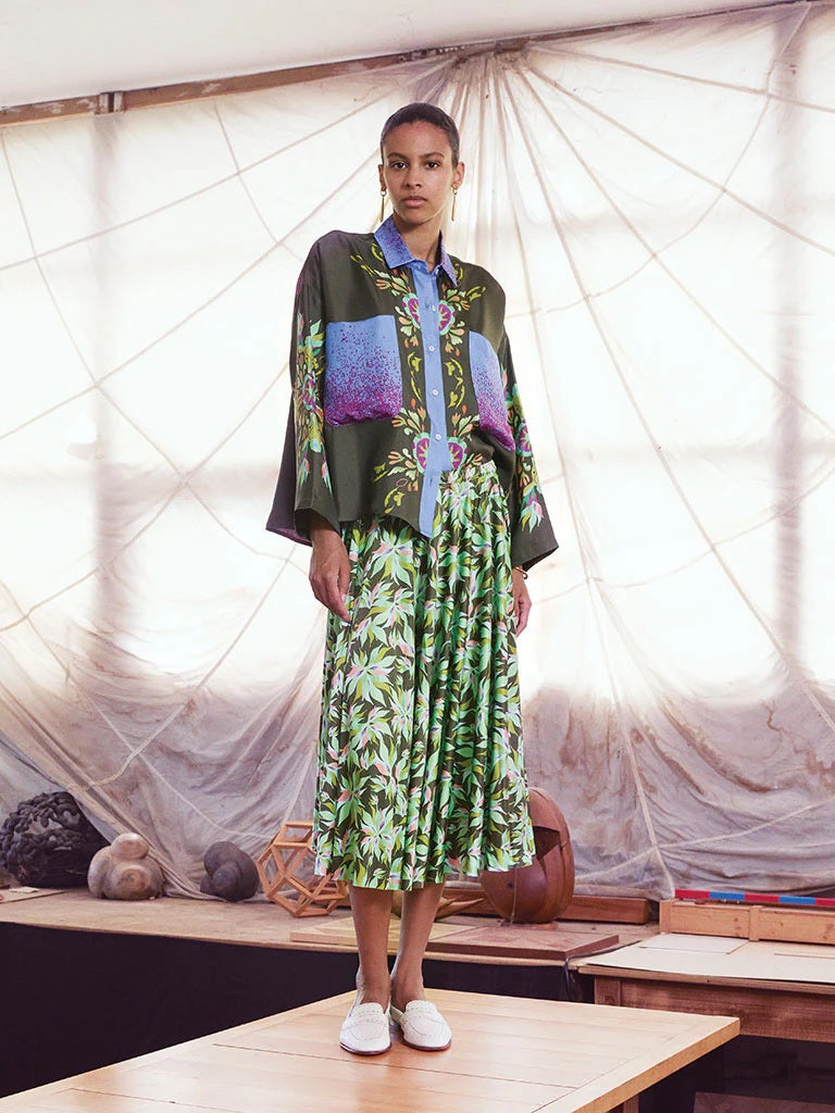 A woman with long brown hair, smiling slightly, wears a loose-fitting, long-sleeve top with colorful patterns, the FRANCO FERRARI Floral Print Skirt from Italy in green, and beige sandals.