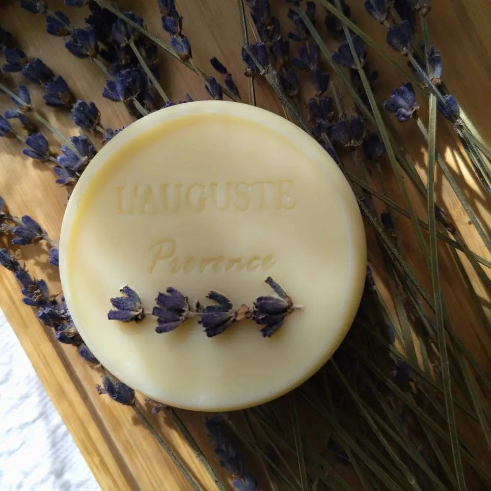 L&#39;AUGUSTE PROVENCE – THE ESSENCE OF PROVENCE Lavender Soap, a round handmade French soap wrapped in cloth with a lavender field and tree motif, rests in an open L&#39;AUGUSTE PROVENCE gift box beside its lid and scattered lavender buds.