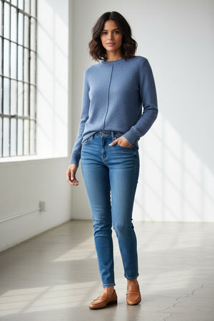 A woman with long, light brown hair wears the Kinross Cashmere Hi Low Seamed Crew Neck Cashmere Sweater in Loch, styled in a relaxed classic fit with blue jeans. She stands against a plain light background, smiling slightly at the camera.