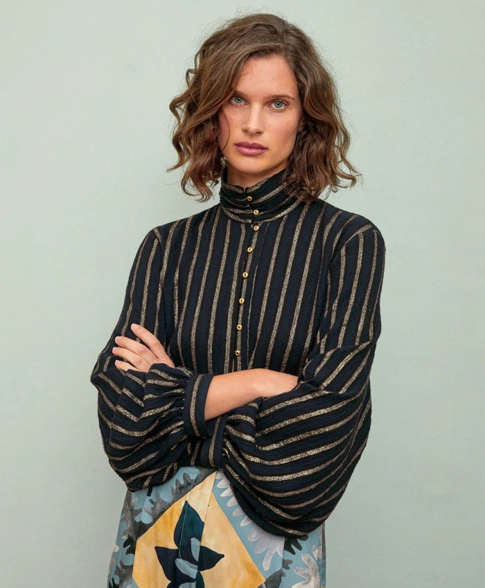 A woman with wavy brown hair, arms crossed, wears the MOMONI Thierry Blouse in lurex stripe gauze and a patterned skirt, standing against a pale background.