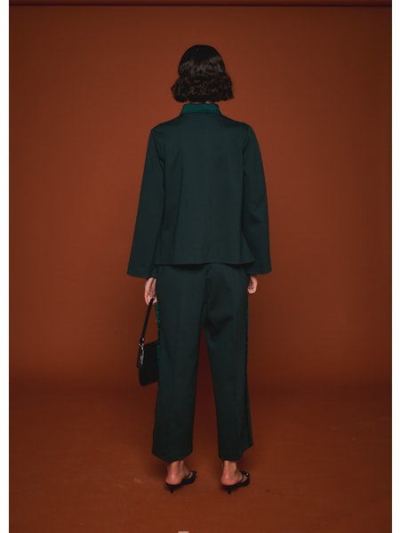Facing away from the camera, a person wears MILVA’s MILVA MI - TUXEDO DRESS PANT in dark green with cropped legs and black heels, holding a black handbag. The background is solid brown.