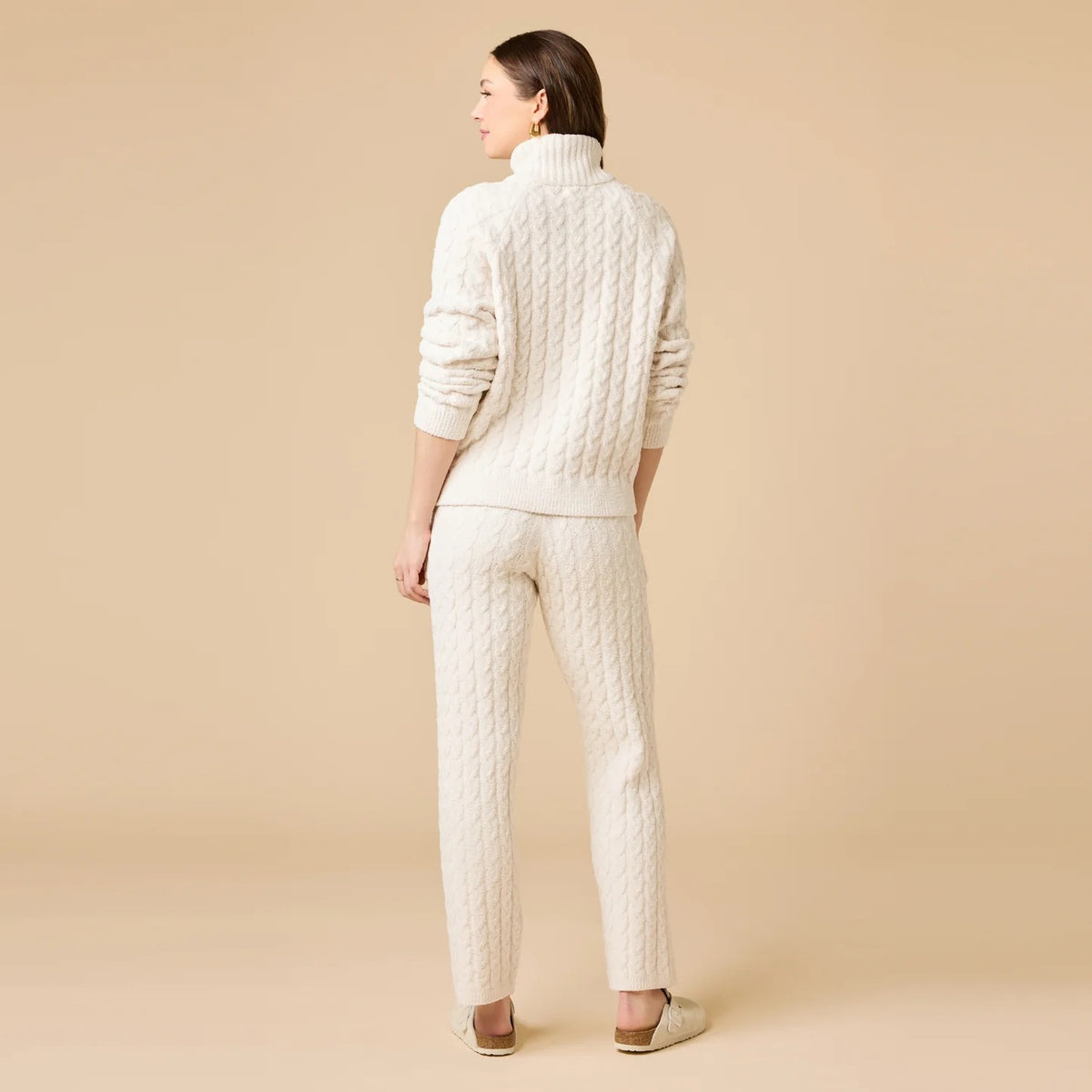 A woman wearing a marshmallow-soft, cream-colored SOFTIES - CABLE KNIT MARSHMALLOW ½ ZIP WITH TAPERED PANT SET, featuring a half-zip pullover and matching pants with a drawstring waist.