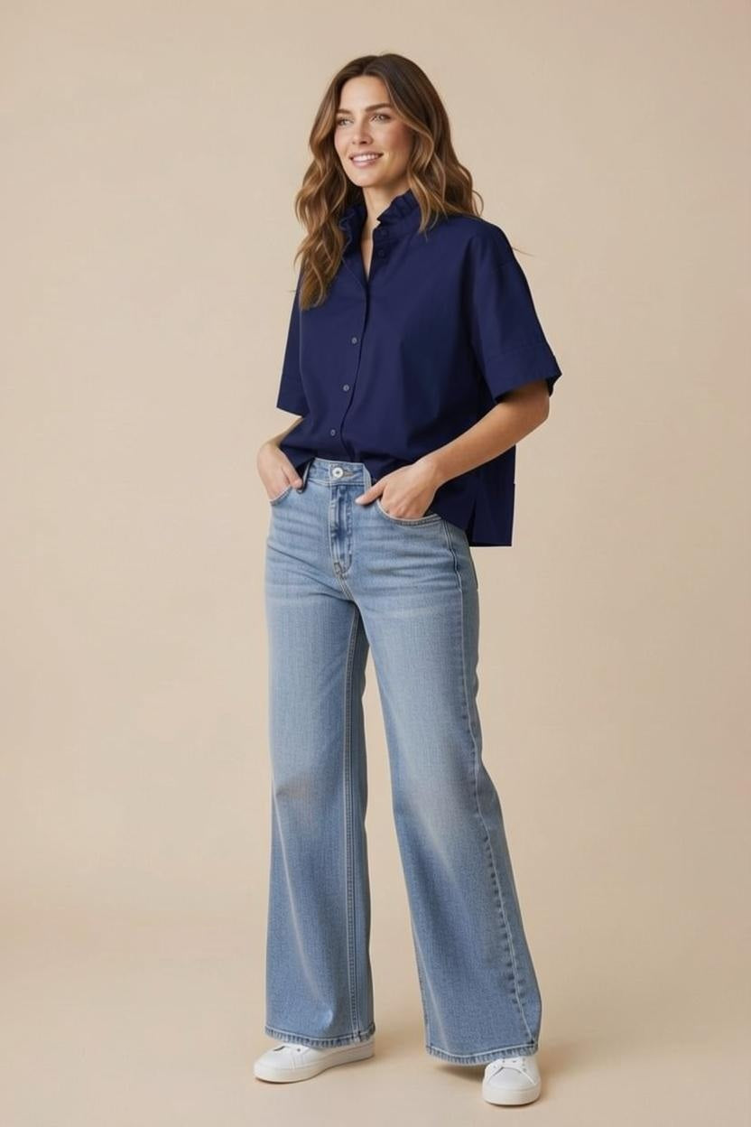 A woman stands smiling with her hands in her pockets against a beige background, wearing the KMJ KMY - REESE SHIRT IN NAVY—a relaxed-fit, short-sleeve button-up—paired with light blue wide-leg jeans and white sneakers.