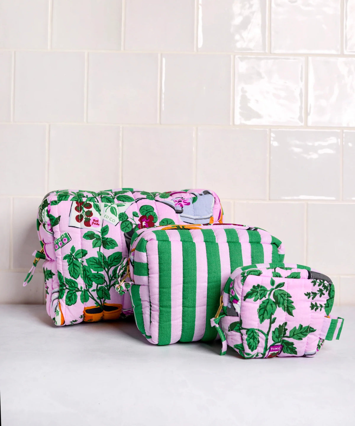 A person in a white shirt holds the PRINTFRESH - GREEN THUMB THREE POUCH SET by PRINTFRESH, featuring three quilted travel pouches with floral and striped patterns, stacked from largest to smallest, against a plain background.