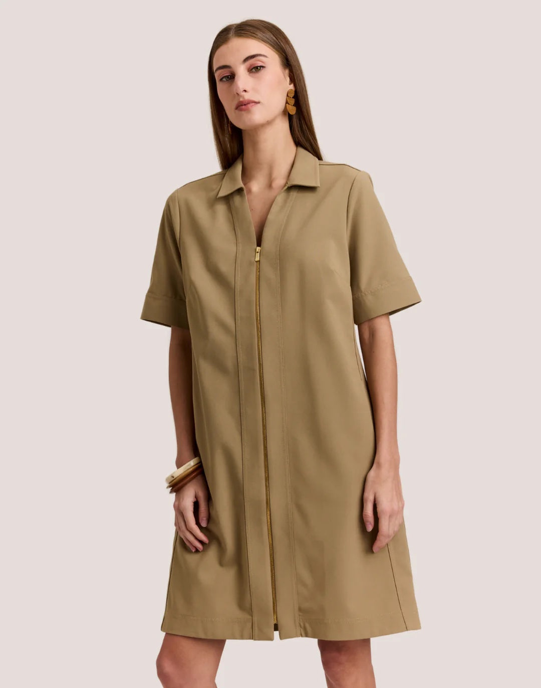 A woman faces forward wearing the HINSON WU BRIDGET Short Sleeve Dress in Traveler’s Twill Khaki, featuring a gold front zipper and one hand in her pocket against a plain light background.