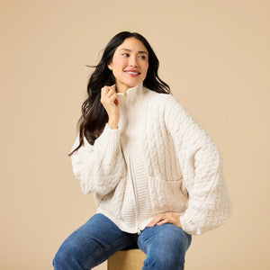 A woman wearing a cream-colored, plush SOFTIES - CABLE KNIT MARSHMALLOW FUNNEL NECK DOUBLE ZIP JACKET with a funnel neck and double zipper, paired with blue jeans.