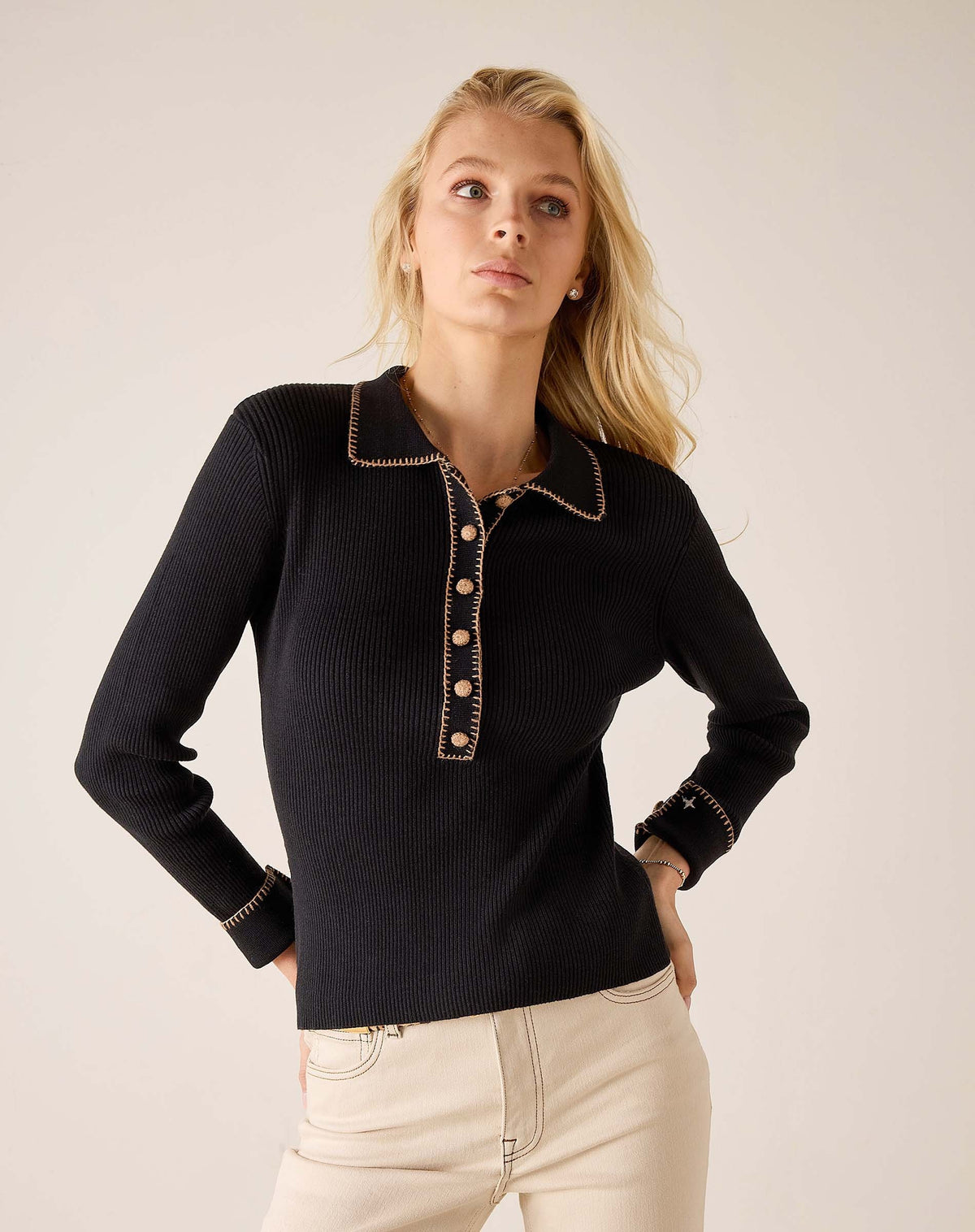 A person with curly hair stands barefoot indoors, smiling near a large window while wearing the MERSEA LUDLOW Long Sleeve Polo in Ink and light-colored jeans.