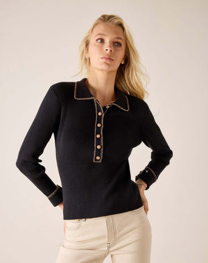 A person with curly hair stands barefoot indoors, smiling near a large window while wearing the MERSEA LUDLOW Long Sleeve Polo in Ink and light-colored jeans.