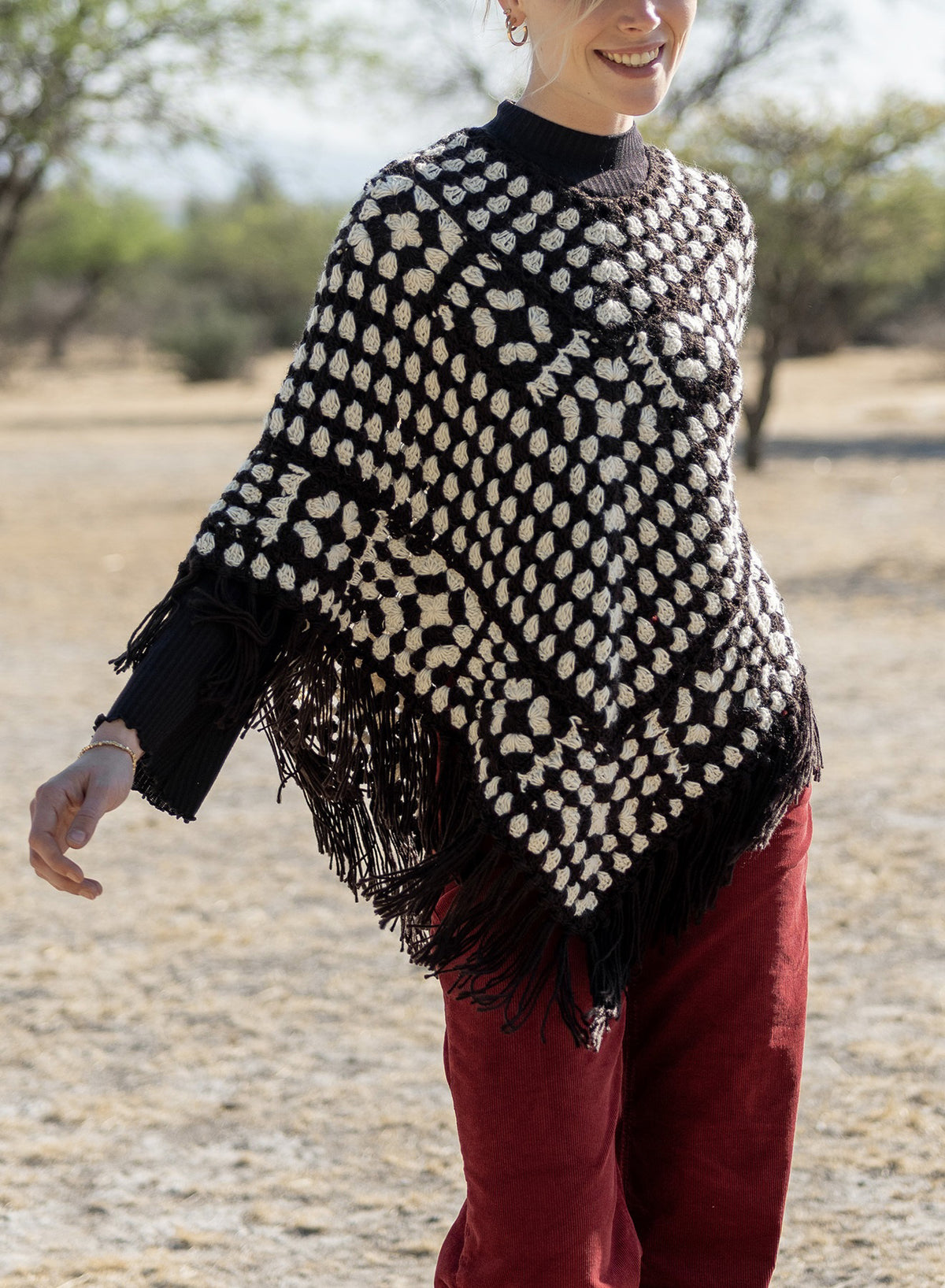 A person outdoors smiles while wearing the MERSEA LA PAZ CROCHET PONCHO—a black and white fringed style by MERSEA—over a dark sweater and red pants, adding bohemian flair amid dry grass and trees.