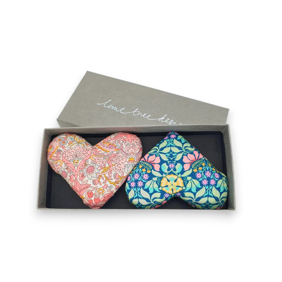 A gray rectangular box with a slightly open lid reveals two heart-shaped items inside. One heart features a pink floral pattern, while the other showcases a blue floral pattern. The phrase &quot;lime tree designs&quot; is elegantly inscribed on the lid, making it an ideal handmade gift from the HEARTS AND MOTHERS PRIDE SACHET collection by LIME TREE COLLECTION LTD.