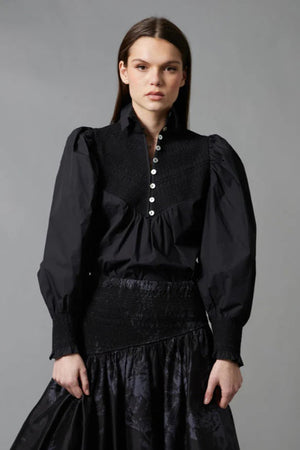 A woman with long brown hair faces away, wearing the HOPE FOR FLOWERS Smocked Shirt in Black with puffed sleeves and paired with a dark floral skirt, standing before a gray backdrop.