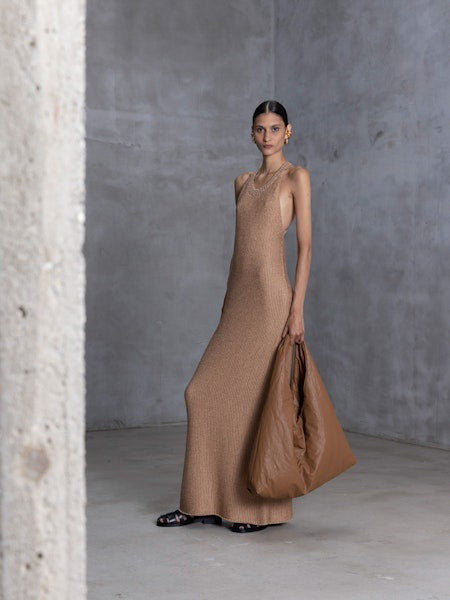 A person in a long, leopard-print outfit stands against a gray wall, holding the NUDE - OVERSIZED HOBO BAG. Their hair is pulled back and they are wearing black sandals. The minimalist design of the concrete floor creates a neutral setting ideal for daily use.